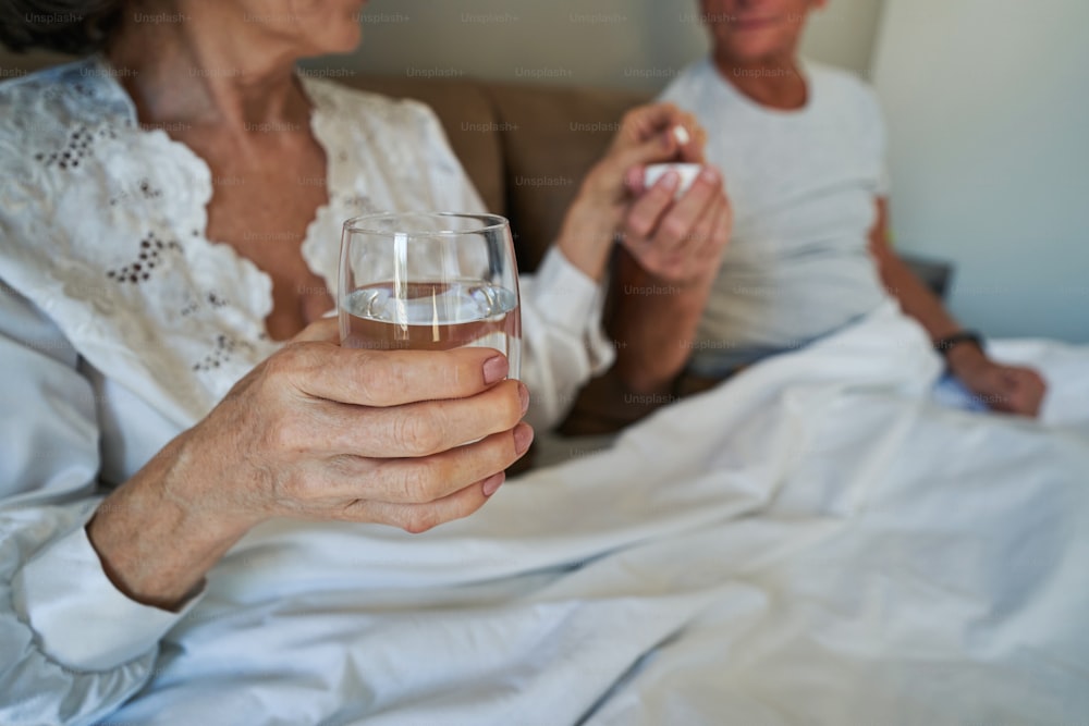 Aging female keeping glass of water in one hand while grabbing medicine from husband with another