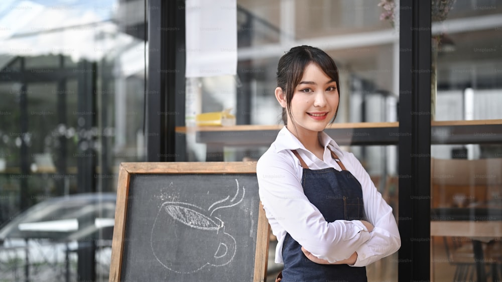 喫茶店の戸口で腕を組んで立っている微笑む若い女性。