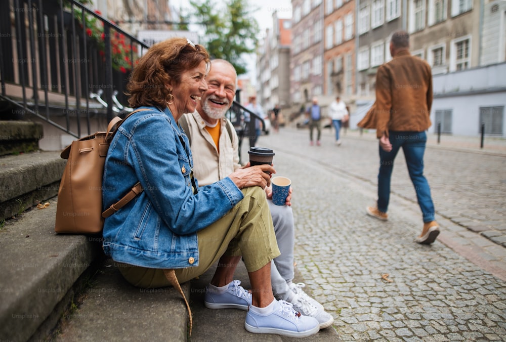 Un heureux couple de touristes âgés assis sur les escaliers et prenant un café à emporter à l’extérieur en ville