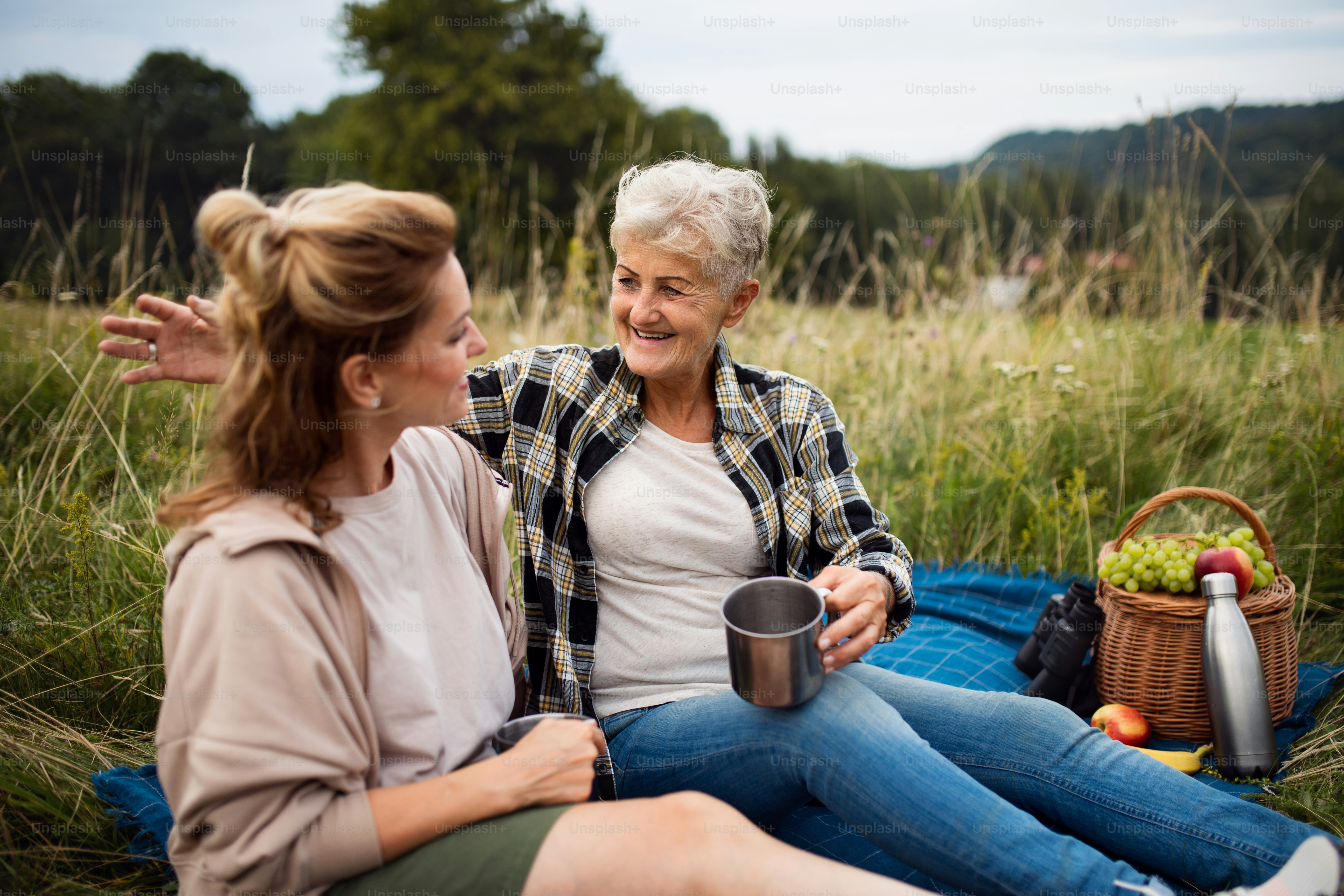Una feliz madre mayor y una hija adulta sentadas y haciendo un picnic al aire libre en la naturaleza, hablando.