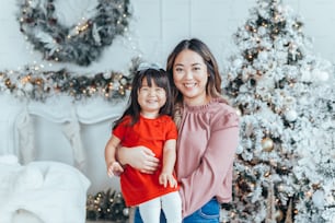 Asian family mother holding daughter toddler girl celebrating Christmas or New Year. Mixed race mom with kid child standing by Christmas tree home. Winter holiday celebration.