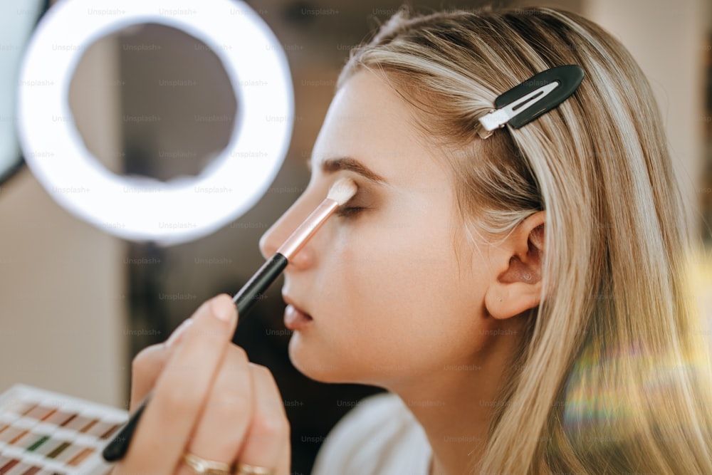 Makeup process. Professional artist applying make up on model face. Close up portrait of beautiful blonde woman in beauty saloon.