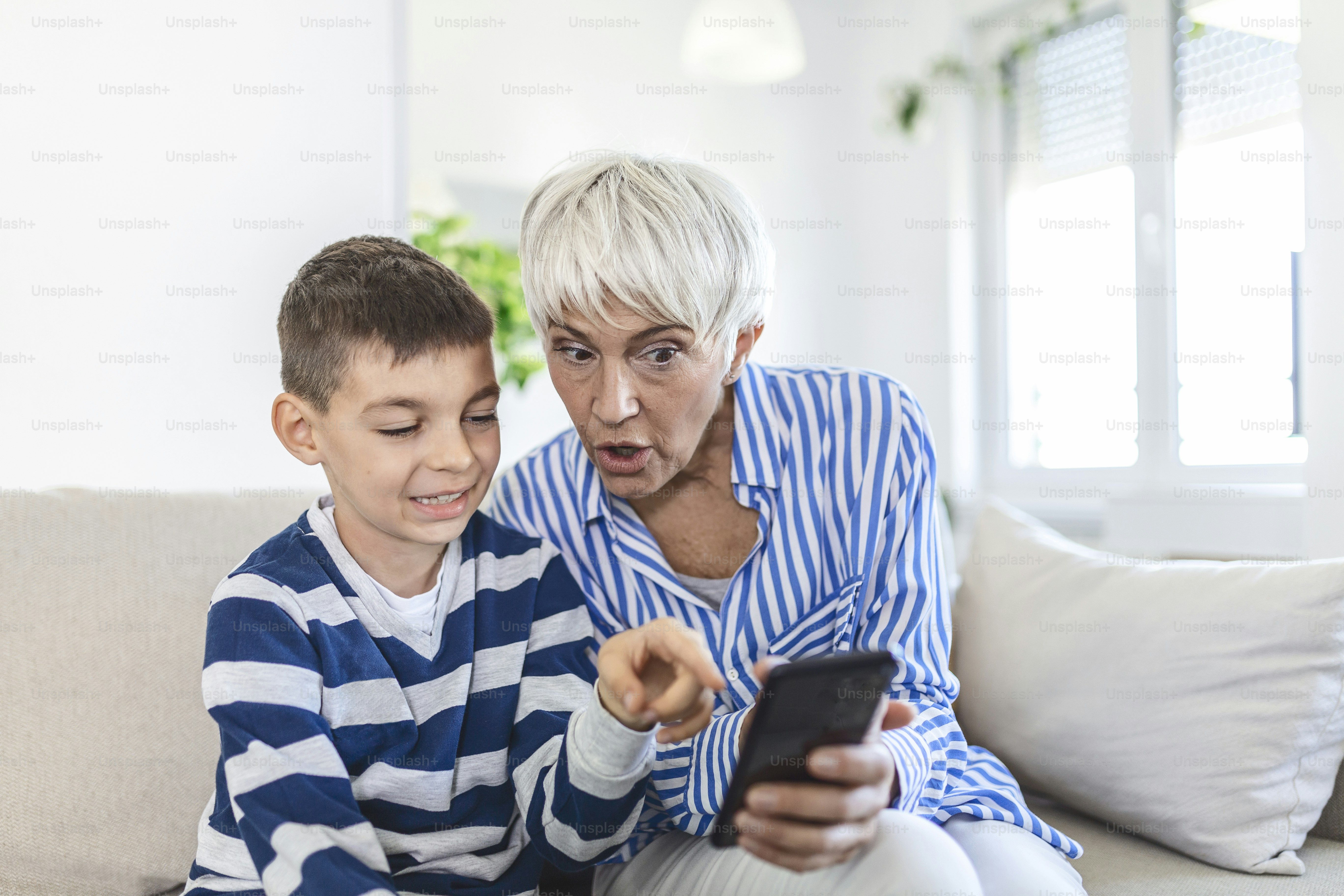 Happy grandmother and grandson using phone together, sitting on cozy sofa at home, browsing mobile device apps, grandma with grandchild playing game, looking at screen, having fun