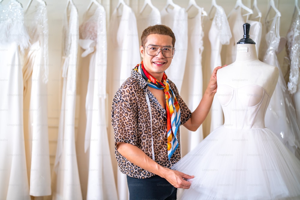 Portrait of Asian LGBTQ guy bridal shop owner using tape measure measuring wedding dress on sewing mannequin in fitting room at wedding studio. Small business entrepreneur wedding planner and tailor designer concept