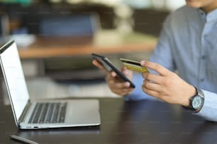 Imagen recortada de un hombre o hombre de negocios sosteniendo una tarjeta de crédito y un teléfono inteligente en su escritorio. Concepto de banca en línea por Internet.