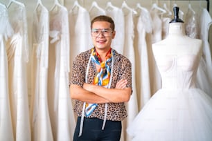 Portrait of Asian LGBTQ guy bridal shop owner using tape measure measuring wedding dress on sewing mannequin in fitting room at wedding studio. Small business entrepreneur wedding planner and tailor designer concept