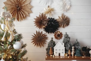 Merry Christmas! Stylish christmas little house, trees, golden lights on background of paper stars on white wooden wall. Atmospheric festive  decor in modern scandinavian room. Magic winter time.