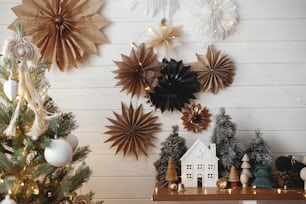 ¡Feliz Navidad! Elegante casita de navidad, árboles, luces doradas sobre fondo de estrellas de papel en pared de madera blanca. Decoración festiva atmosférica en una habitación escandinava moderna. Mágico horario de invierno.