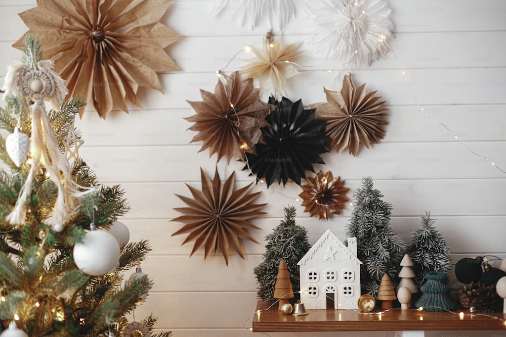 Merry Christmas! Stylish christmas little house, trees, golden lights on background of paper stars on white wooden wall. Atmospheric festive  decor in modern scandinavian room. Magic winter time.