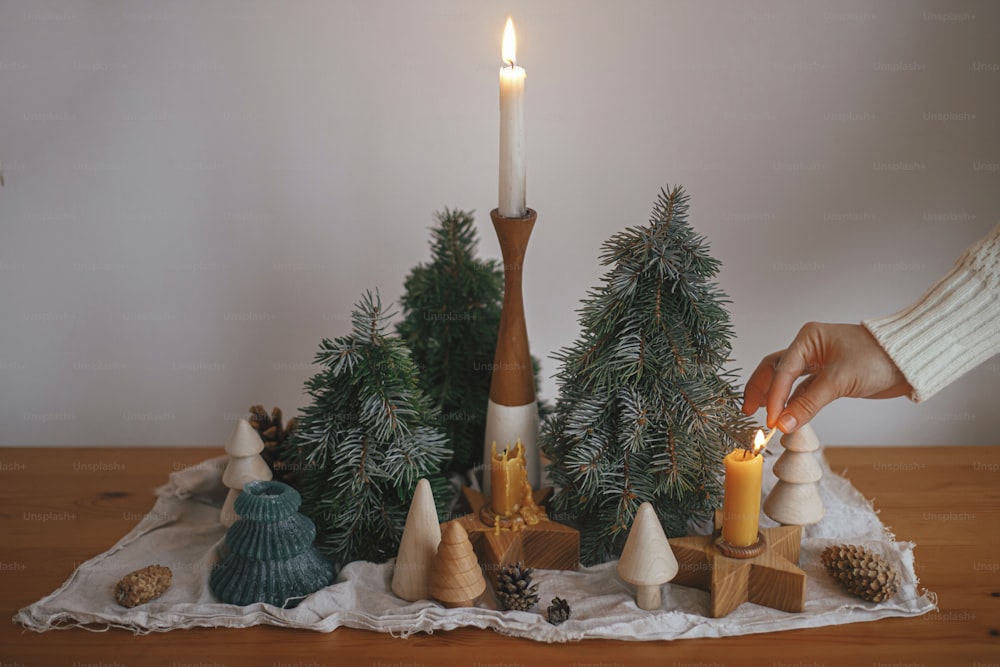 Feiertag Advent. Hand in kuscheligem Pullover Beleuchtung Weihnachtskerze auf dem Hintergrund von Pinien Dekorationen, Zapfen, rustikalen Stoff auf Holztisch im Abend skandinavischen festlichen Raum.  Atmosphärisch