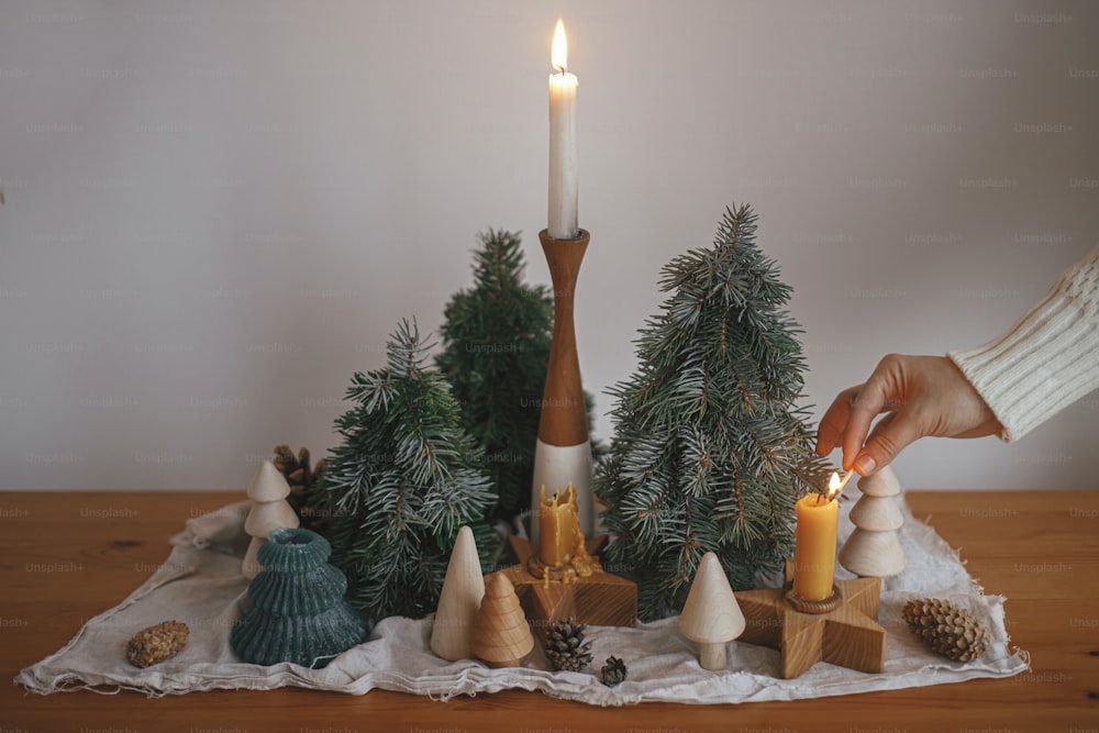 Holiday advent. Hand in cozy sweater lighting up christmas candle on background of pine trees decorations, cones, rustic cloth on wooden table in evening scandinavian festive room.  Atmospheric