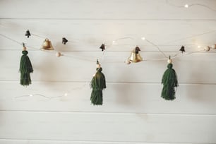 Stylish christmas garland with handmade thread pine trees and bells on white wooden wall with christmas lights. Festive decorated atmospheric scandinavian room. Boho decorations