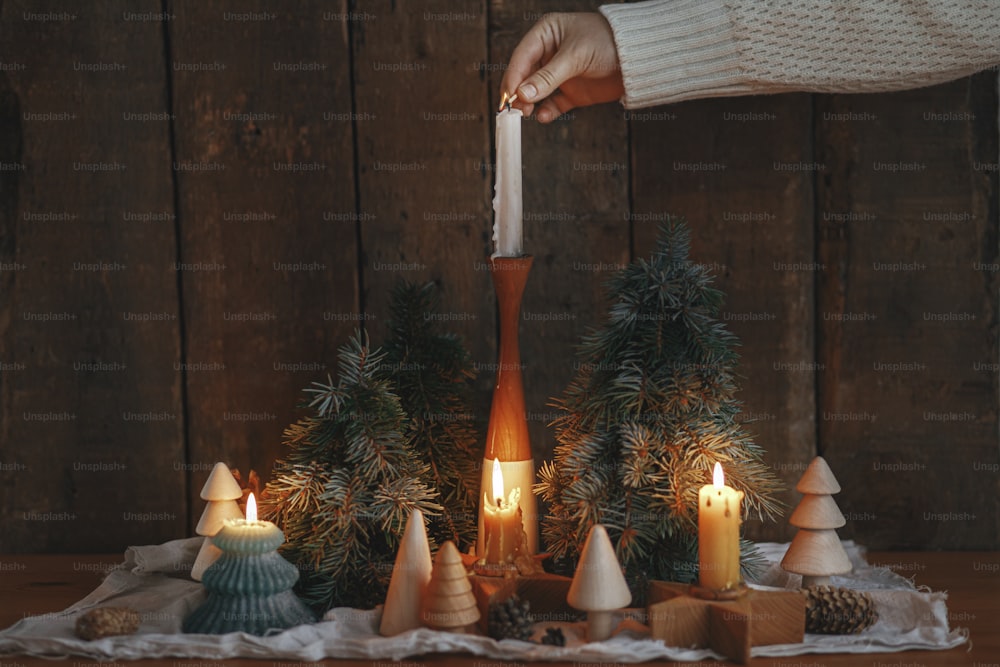 Hand in cozy sweater lighting up christmas candle on rustic wooden background with pine trees and cones in evening scandinavian room. Holiday advent. Atmospheric moment