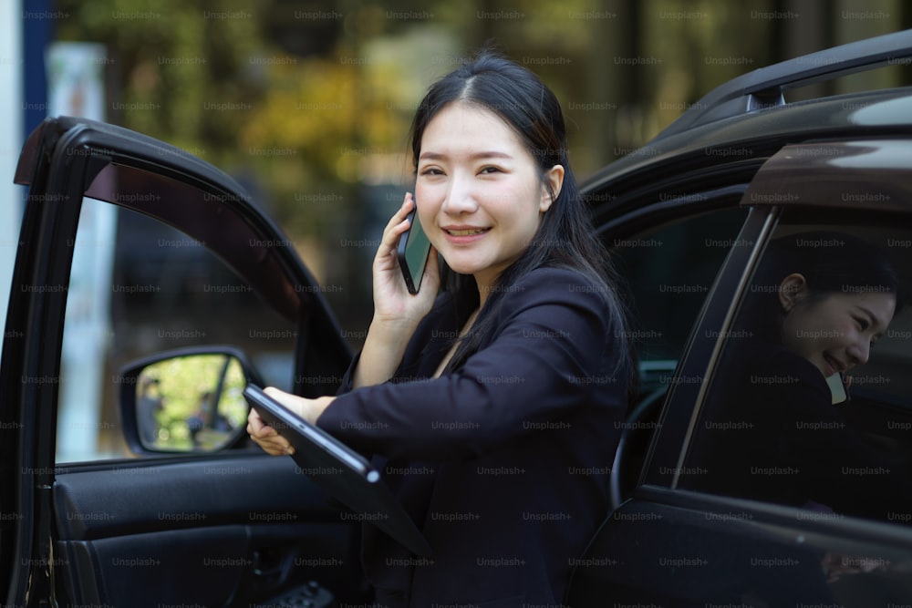 車から降りながら電話でビジネストークをするスマートな働く女性。忙しいビジネスウーマンのコンセプト。