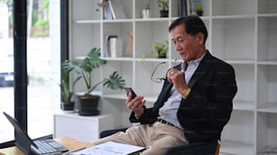 Mature businessman sitting in his office room and using smart phone.