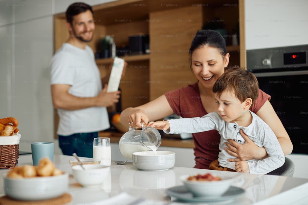 Glückliche Mutter und Sohn gießen Milch in eine Schüssel, während sie am Esstisch frühstücken. Vater ist im Hintergrund.