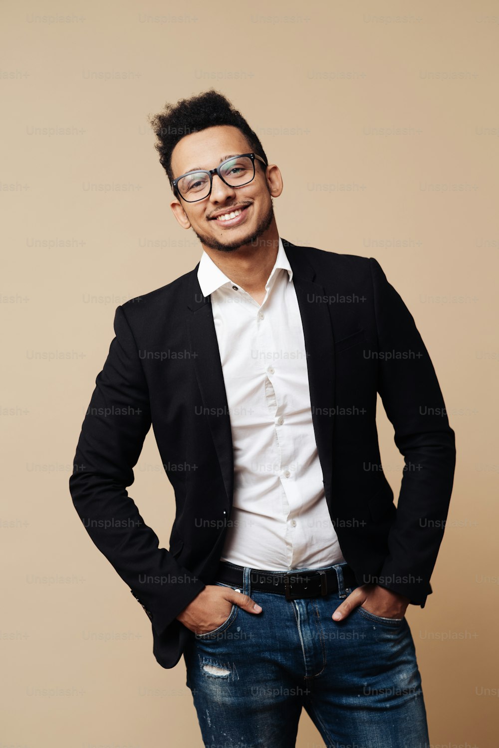Portrait of young Afro businessman wearing shirt, black suit, glasses and looking at camera while standing at beige background.
