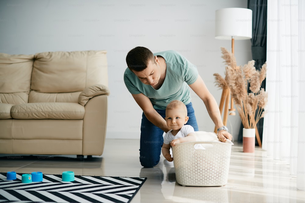 Small boy sitting in laundry basket while father is pushing him in the living room. Copy space.