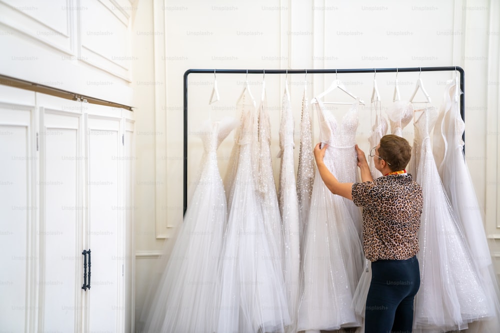 アジアのLGBTQの男のブライダルショップのオーナーは、ウェディングスタジオの試着室で女性の花嫁の顧客のために洋服ラックにぶら下がっているウェディングドレスを選んでいます。中小企業の起業家、ウェディングプランナー、テーラーデザイナーのコンセプト