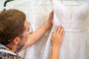 Propriétaire d’une boutique de mariage de gars LGBTQ asiatique choisissant une robe de mariée accrochée sur un porte-vêtements pour une cliente mariée dans la cabine d’essayage du studio de mariage. Concept de concepteur de mariage et de concepteur de petites entreprises, d’entrepreneur et de tailleur
