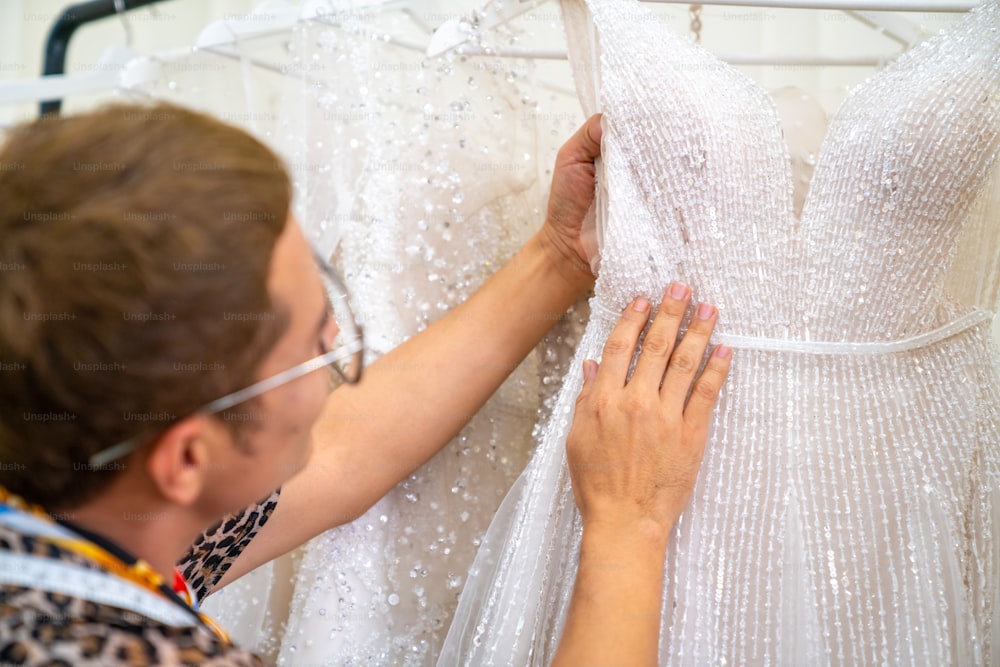 Asian LGBTQ guy bridal shop owner choosing wedding dress hanging on clothes rack for female bride customer in fitting room at wedding studio. Small business entrepreneur wedding planner and tailor designer concept