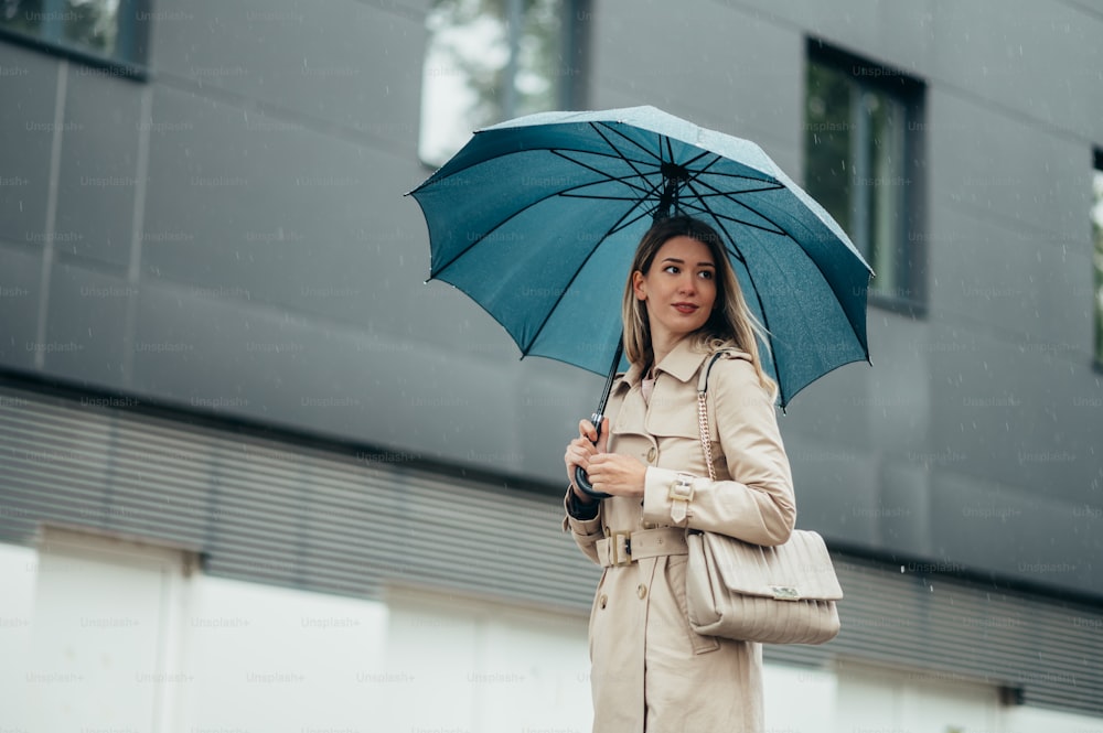 Empresária sorridente jovem com guarda-chuva e bolsa de ombro andando pela rua da cidade durante a chuva