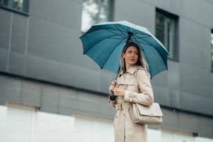雨の中、街の通りを歩いている傘とショルダーバッグを持つ若い笑顔のビジネスウーマン