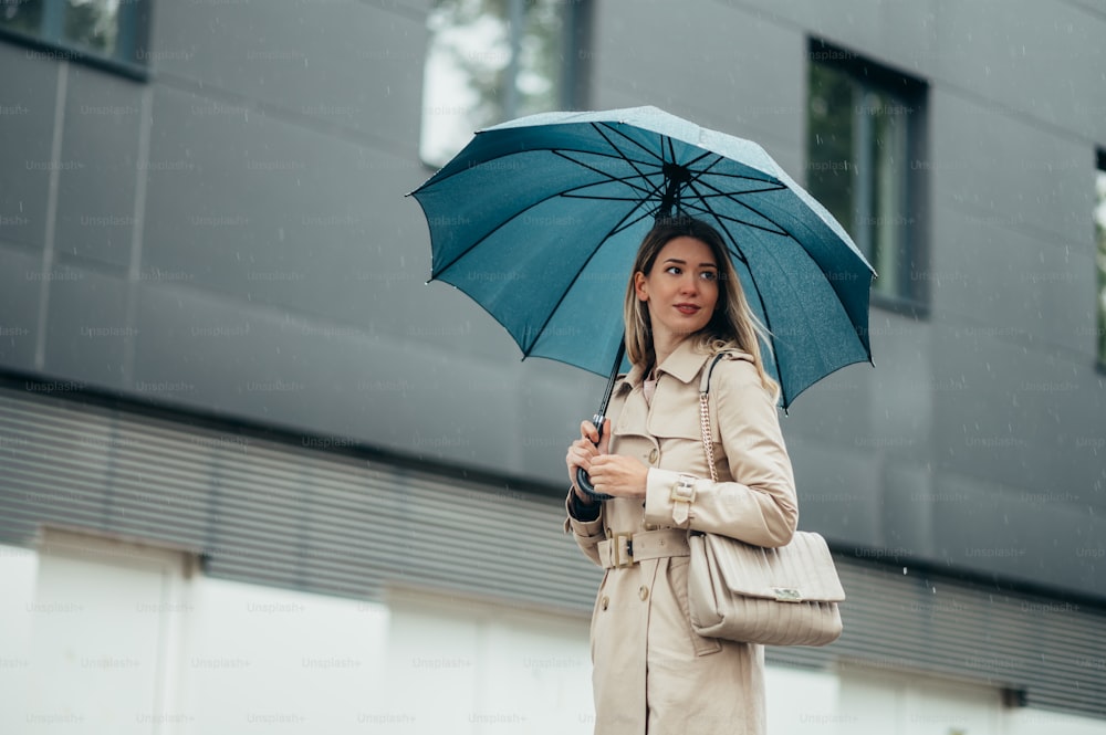Young smiling businesswoman with umbrella and shoulder bag walking down city street during rain