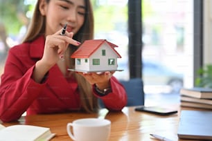 Mujer asiática sonriente sosteniendo un modelo de casa pequeña. Concepto de inversión hipotecaria e inmobiliaria.