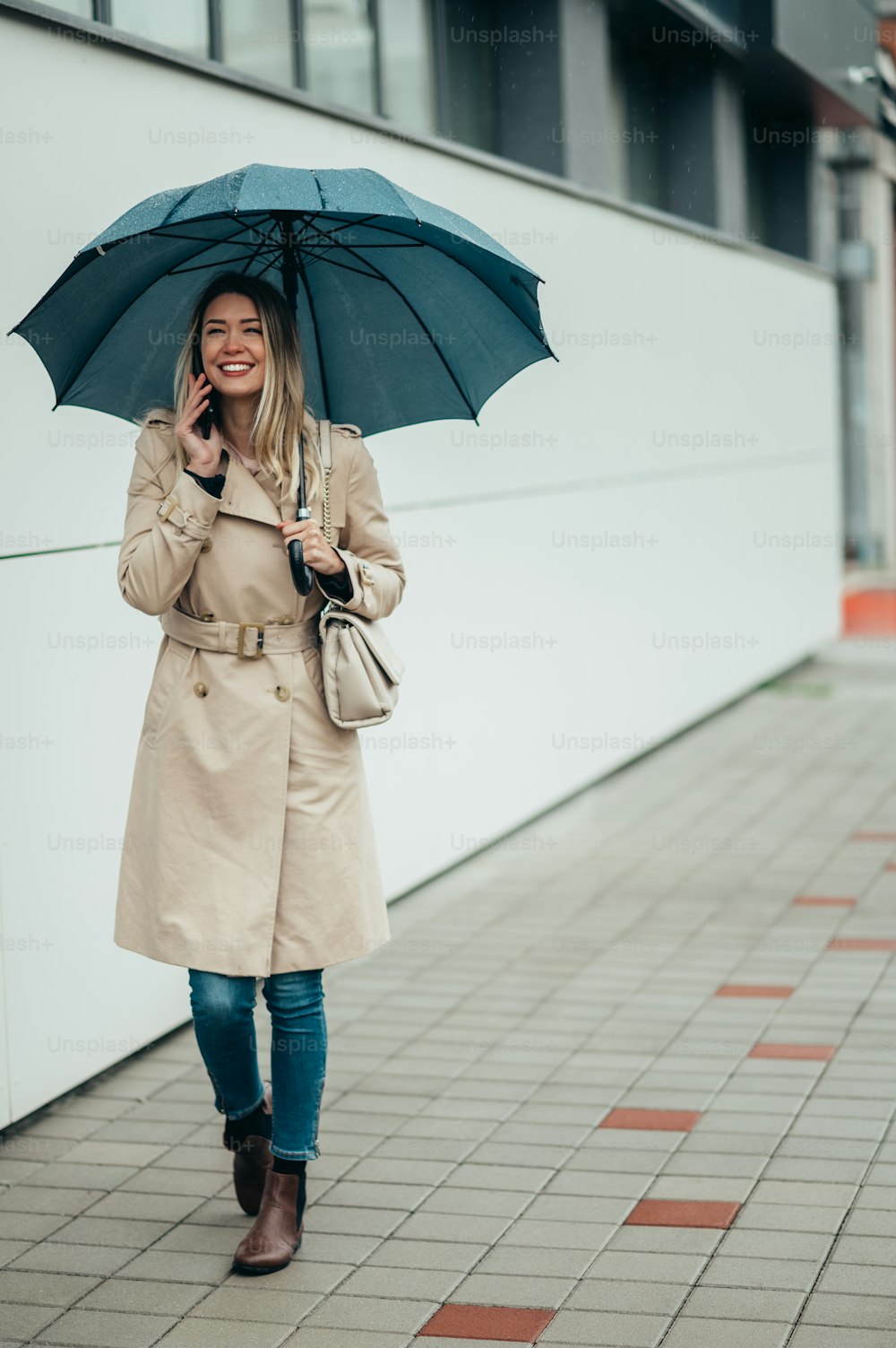 Young smiling businesswoman with umbrella and shoulder bag using smartphone and walking down city street during rain