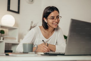 Beautiful woman writing in a notebook and using a laptop while managing her business and working remotely from home