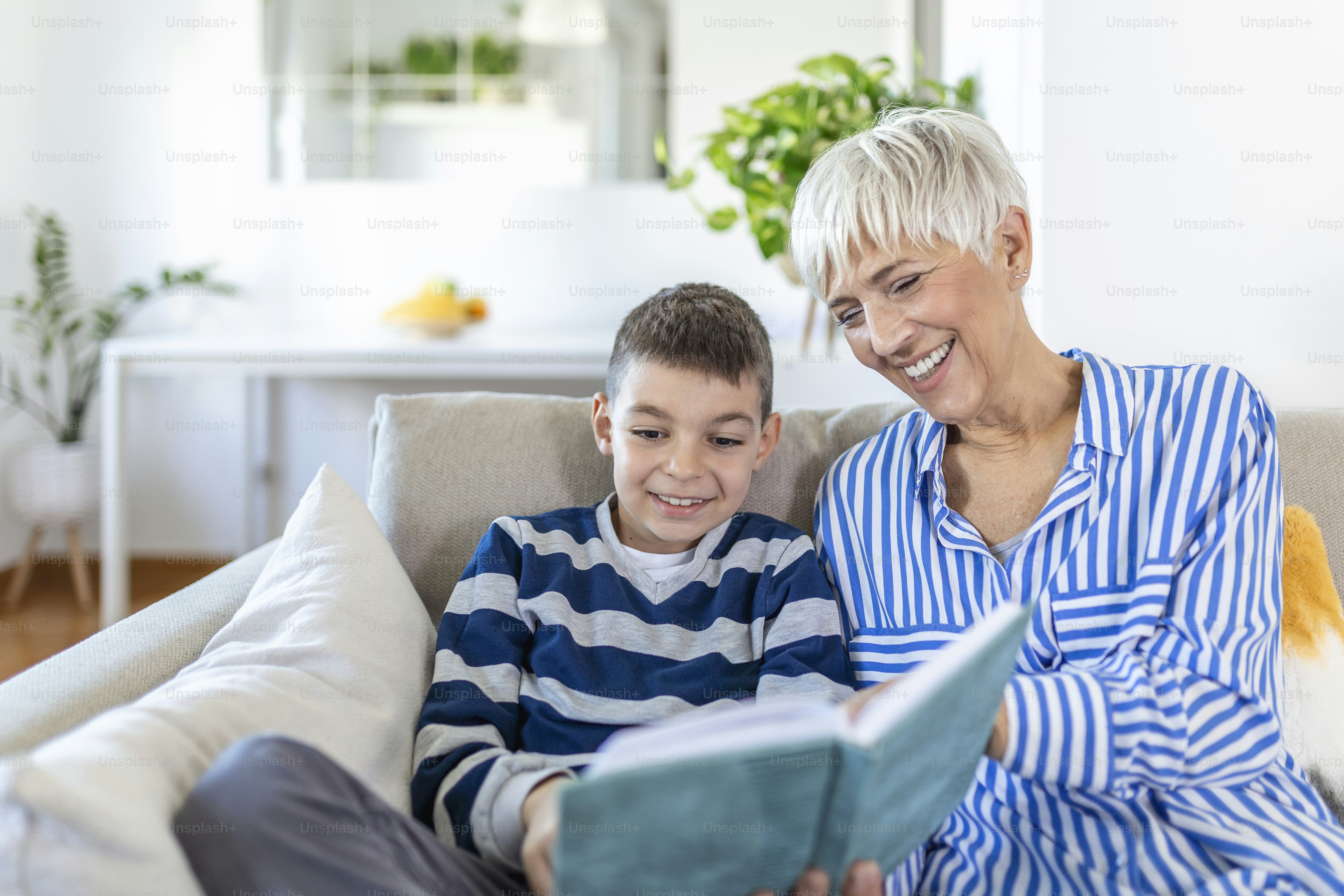 Cropped photo of happy gray-haired woman looking at book with her grandson indoors. They are smiling during their conversation. Visiting grandparents concept