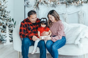 Asian family father, mother giving gift box with present to daughter toddler girl celebrating Christmas or New Year. Mixed race mom, dad, daughter sitting on couch at home. Winter holiday celebration.