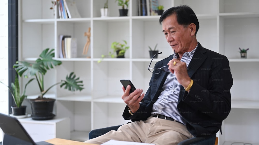Mature businessman holding eyeglasses and using smart phone in office.