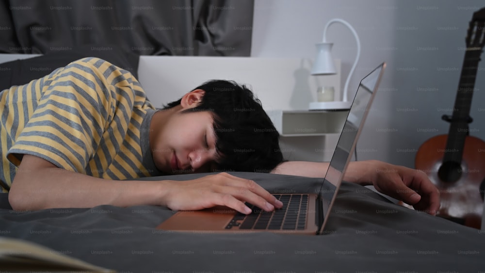 Tired young asian man sleeping on bed near computer laptop.