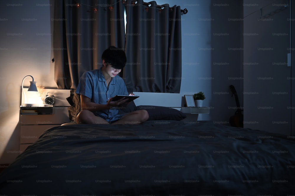 잠옷을 입은 청년이 편안한 침대에 앉아 디지털 태블릿을 사용하고 있다.