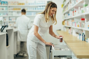 Beautiful female pharmacist working in a pharmacy while checking medications on the shelf