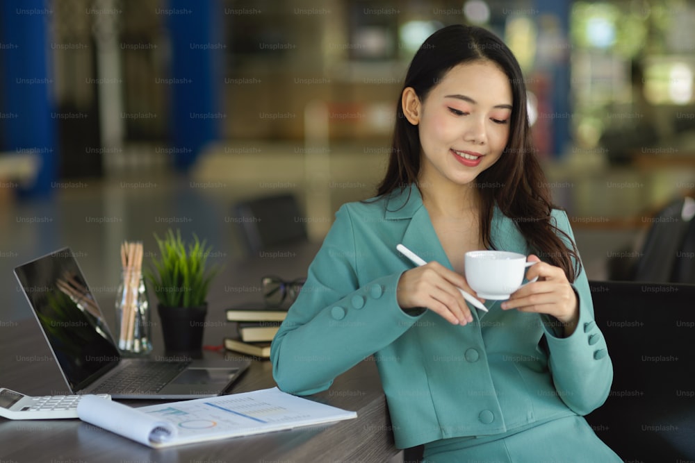 Erfolgreiche asiatische junge Geschäftsfrau oder Managerin, die im Sitzungssaal sitzt und heißen Tee oder Kaffee trinkt.