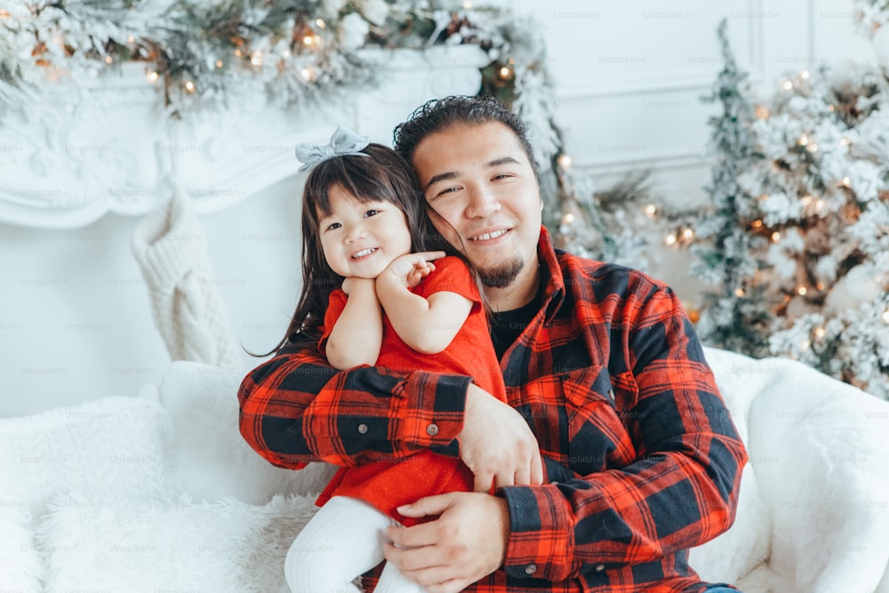 Asian family father dad hugging with daughter toddler girl celebrating Christmas or New Year. Mixed race dad man with kid sitting on couch sofa and cuddling at home. Winter holiday celebration.