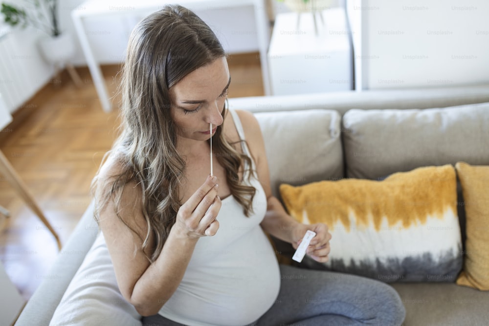Mujer embarazada tomando un autohisopado casero pruebas de COVID-19 en casa con kit de antígenos. Introducción de la punción nasal para controlar la infección por Coronavirus. Cuarentena, pandemia.