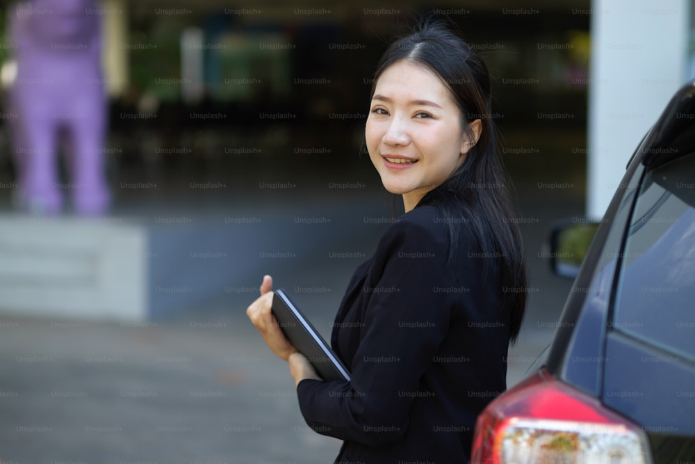A beautiful asian businesswoman in formal suit with tablet computer standing outside near the car.