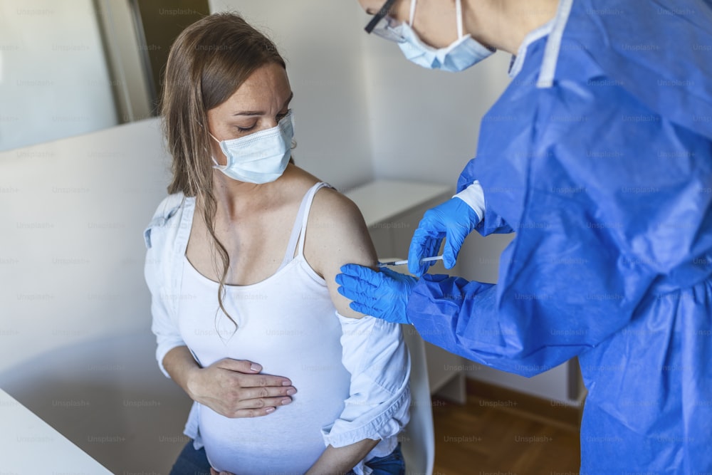 Female doctor in protective face mask and gloves preparing to inject pregnant patient with covid antiviral vaccine, rubbing hand with cotton pad. Mass vaccination centre, global immunization campaign