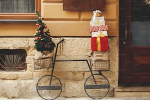 Stylish modern christmas tree with ornaments and pile wrapped gifts on minimalist bicycle at old building. Christmas festive decor for winter holidays in european city street. Merry Christmas!