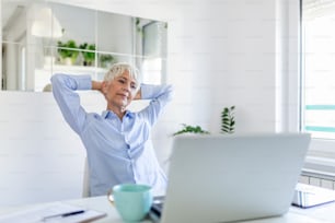 Focused old woman with white hair at home using laptop. Senior stylish entrepreneur working on computer at home. Woman analyzing and managing domestic bills and home finance