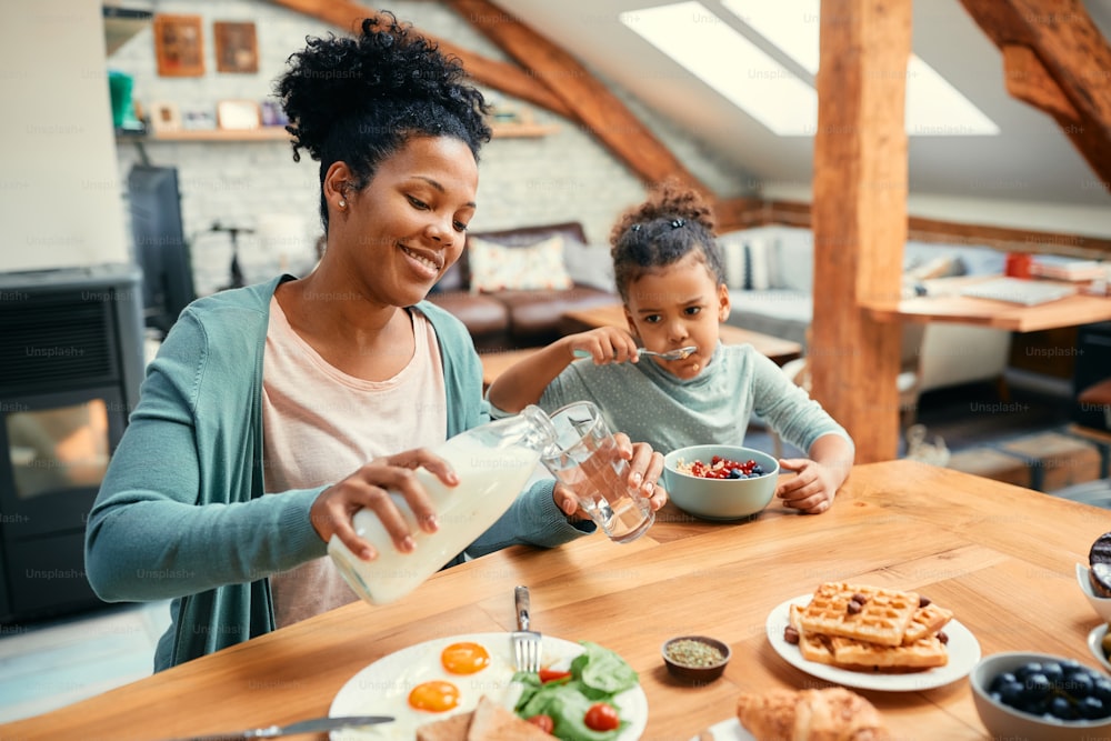 Glückliche afroamerikanische Mutter, die Milch in ein Glas gießt, während sie mit ihrer Tochter am Esstisch frühstückt.