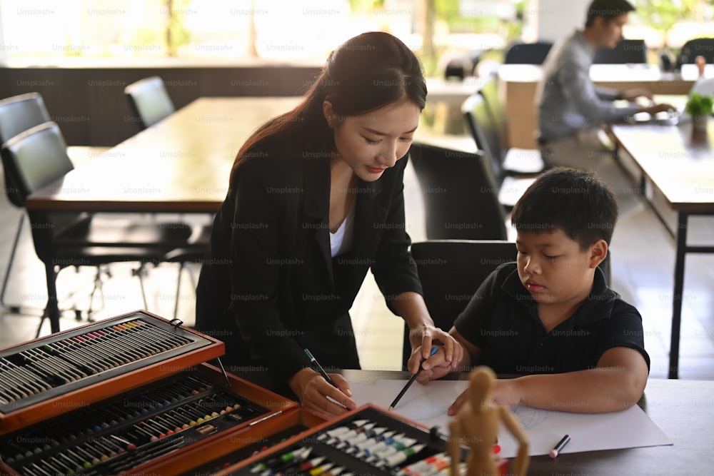 美術の授業で子供たちが絵を着色するのを手伝っている笑顔のアジアの先生。