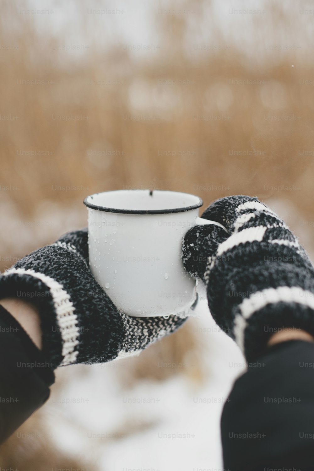 Hands in cozy gloves holding warm cup of tea on background of dried grass and snow lake in winter. Hiking and traveling in cold winter season. Warm drink. Space for text