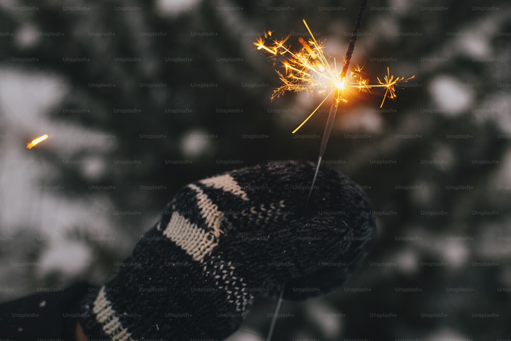 Frohes neues Jahr!! Hand in kuscheligem Handschuh, der brennende Wunderkerze auf dem Hintergrund von Kiefernzweigen im Schnee hält. Atmosphärischer magischer Moment. Frauenhand mit glühendem Feuerwerk am Abend. Frohe Feiertage!