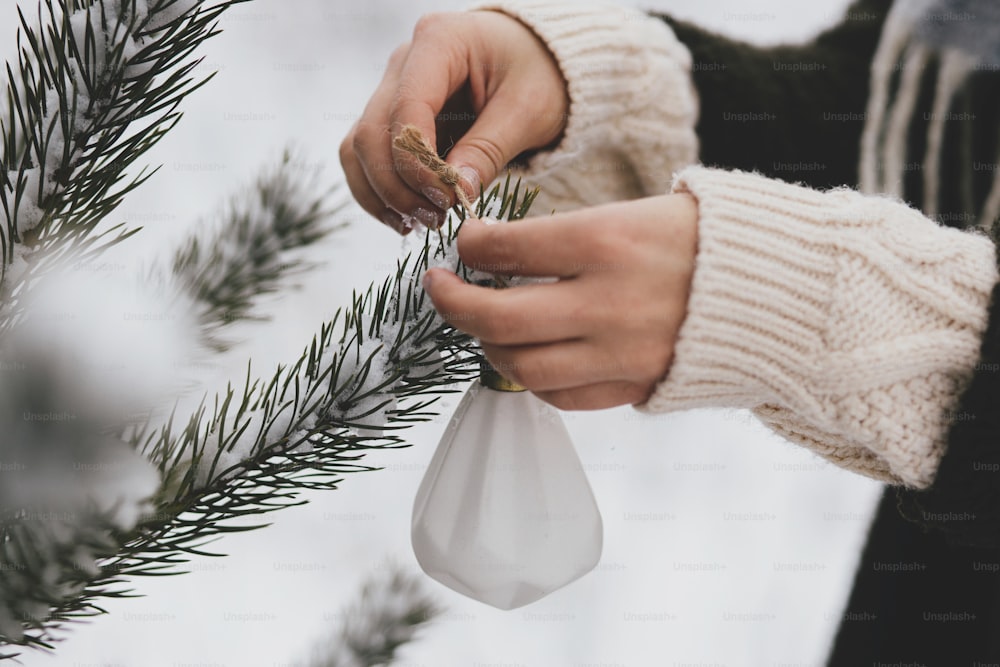 Hands in cozy sweater holding stylish ornament on background of pine tree branches in snow. Decorating christmas tree outdoors.  Preparation for winter holidays in countryside. Merry christmas!