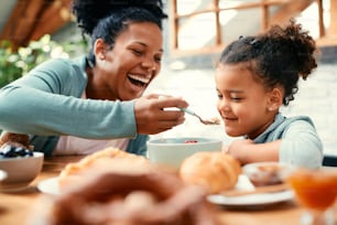 Felice madre nera che si diverte e nutre sua figlia mentre fa colazione al tavolo da pranzo.